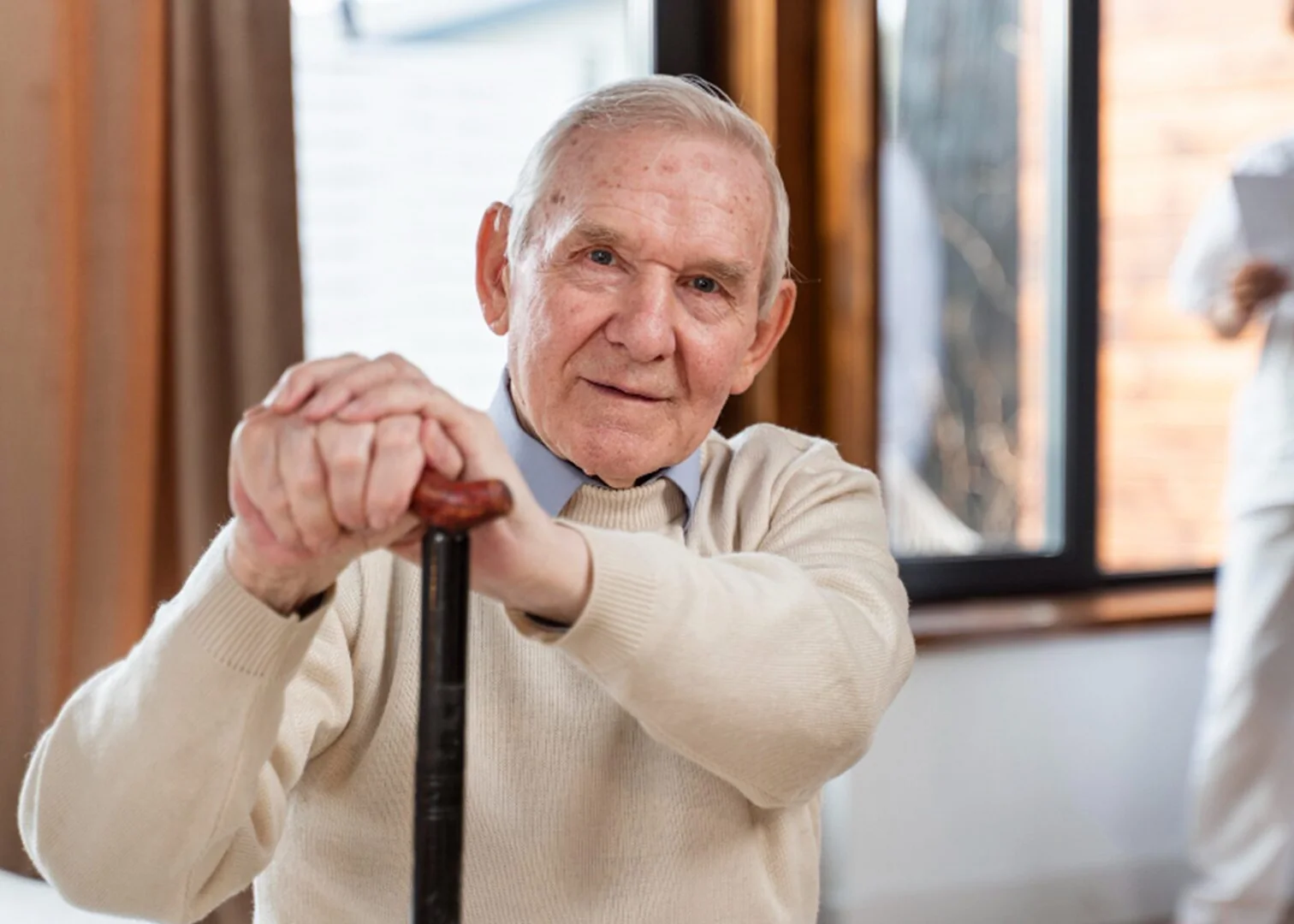 An older man resting on his cane at home while a care professional reviews his notes in the background